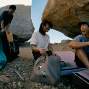 3 climbers sitting and laughing near a boulder with tote bags in the foreground