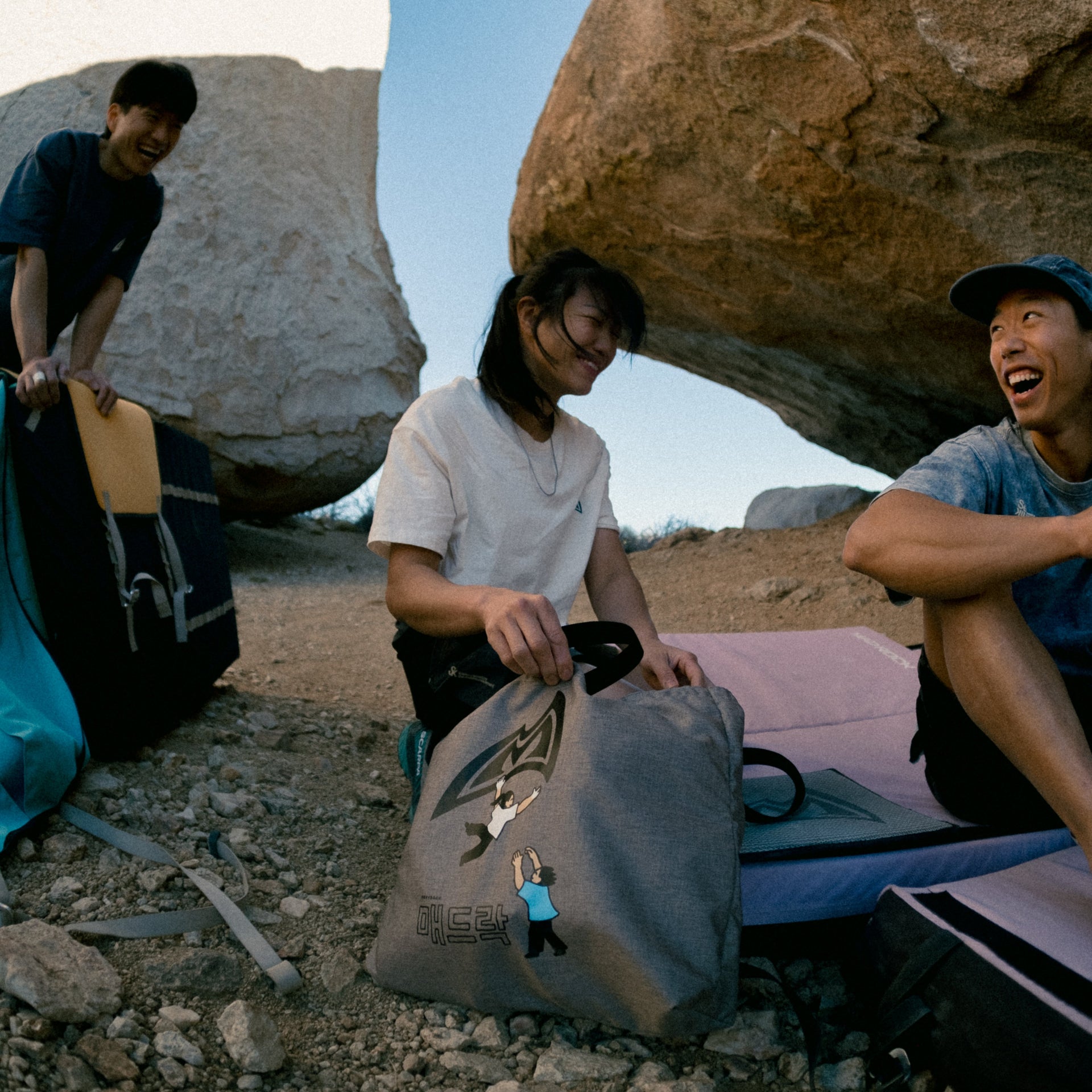 3 climbers sitting and laughing near a boulder with tote bags in the foreground