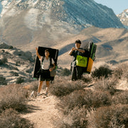 two climbers with crash pads on their backs, carrying totes in a scenic mountain landscape