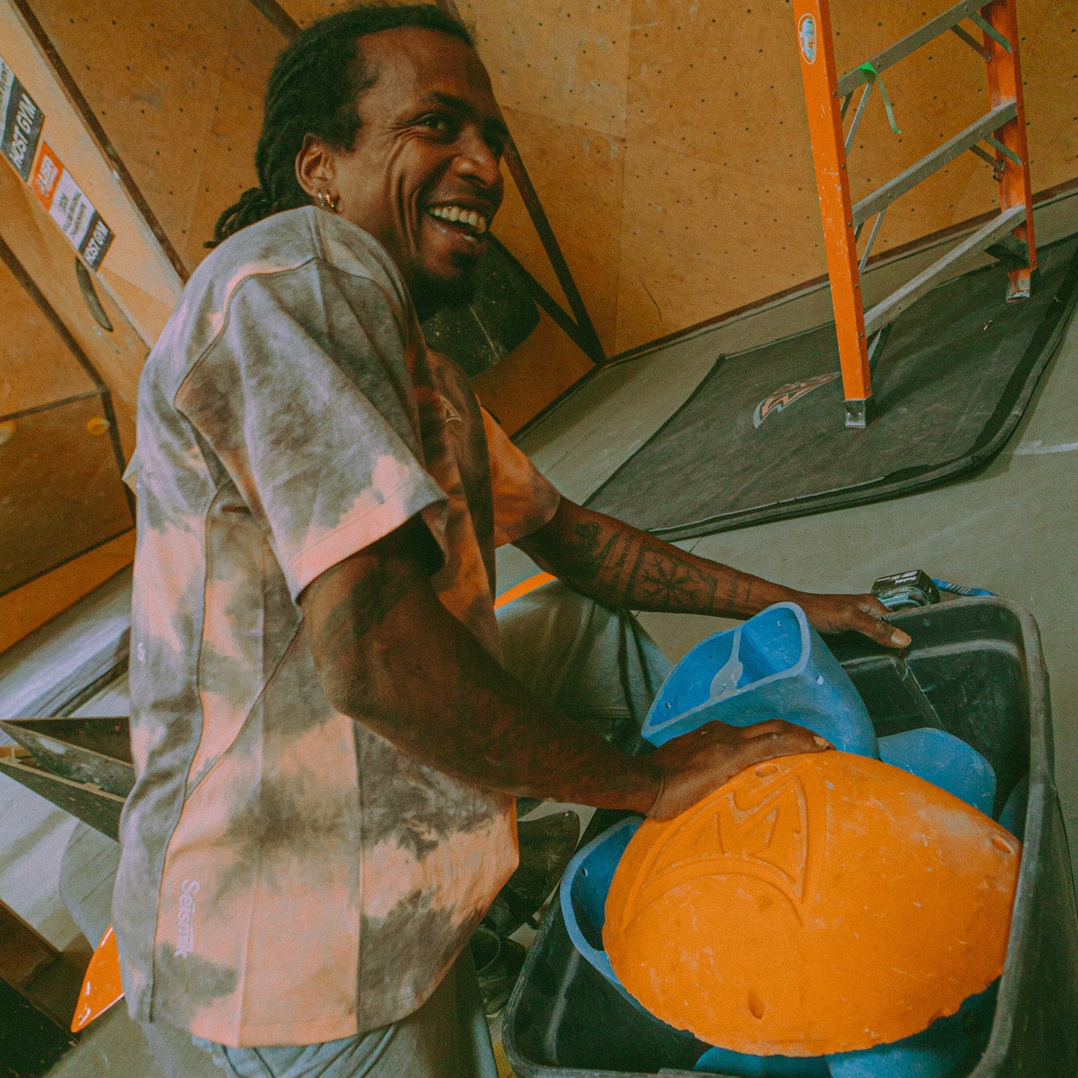 athlete Mickael Mawem wearing an acid wash shirt and happily sifting through climbing holds inside of a plastic tub