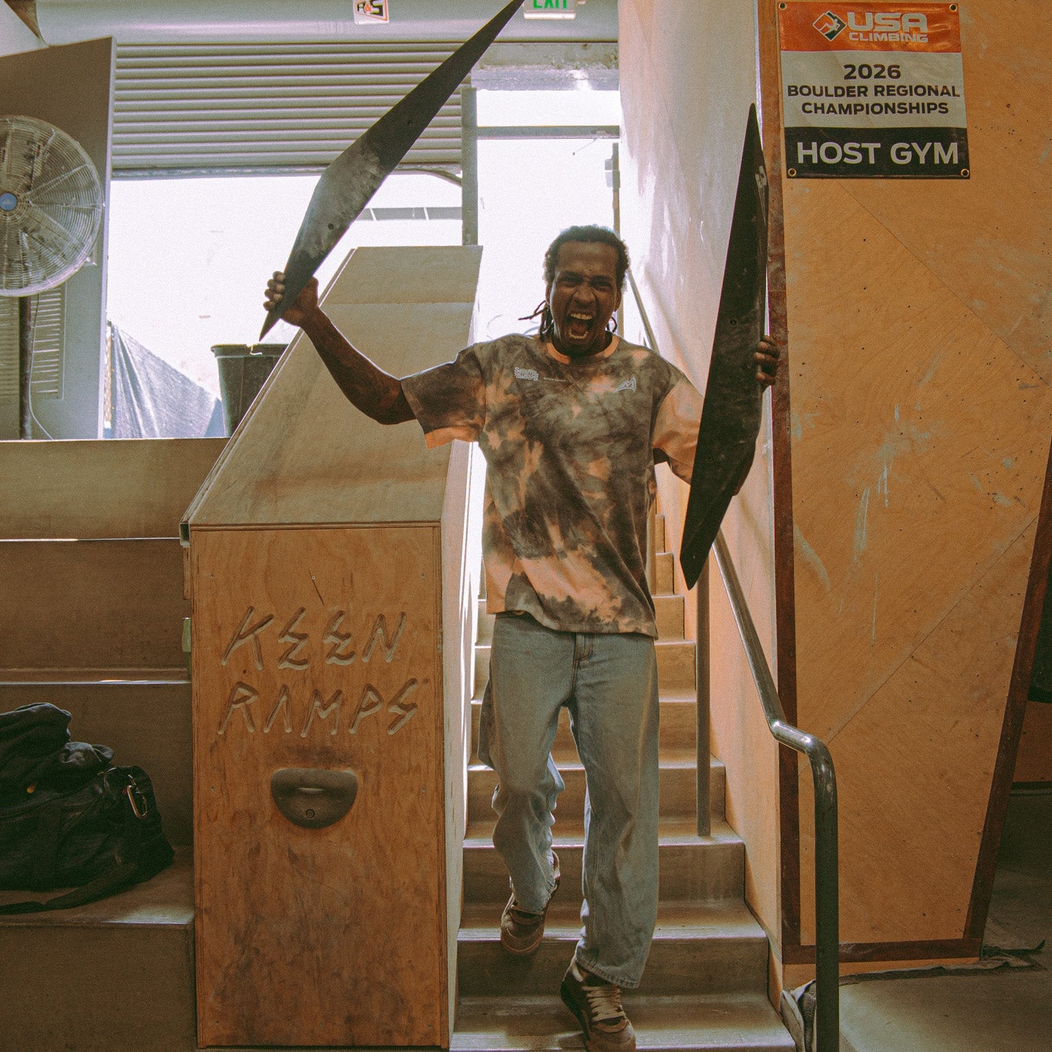 athlete Mickael Mawem wearing an acid wash shirt while carrying two large climbing holds and shouting at the camera