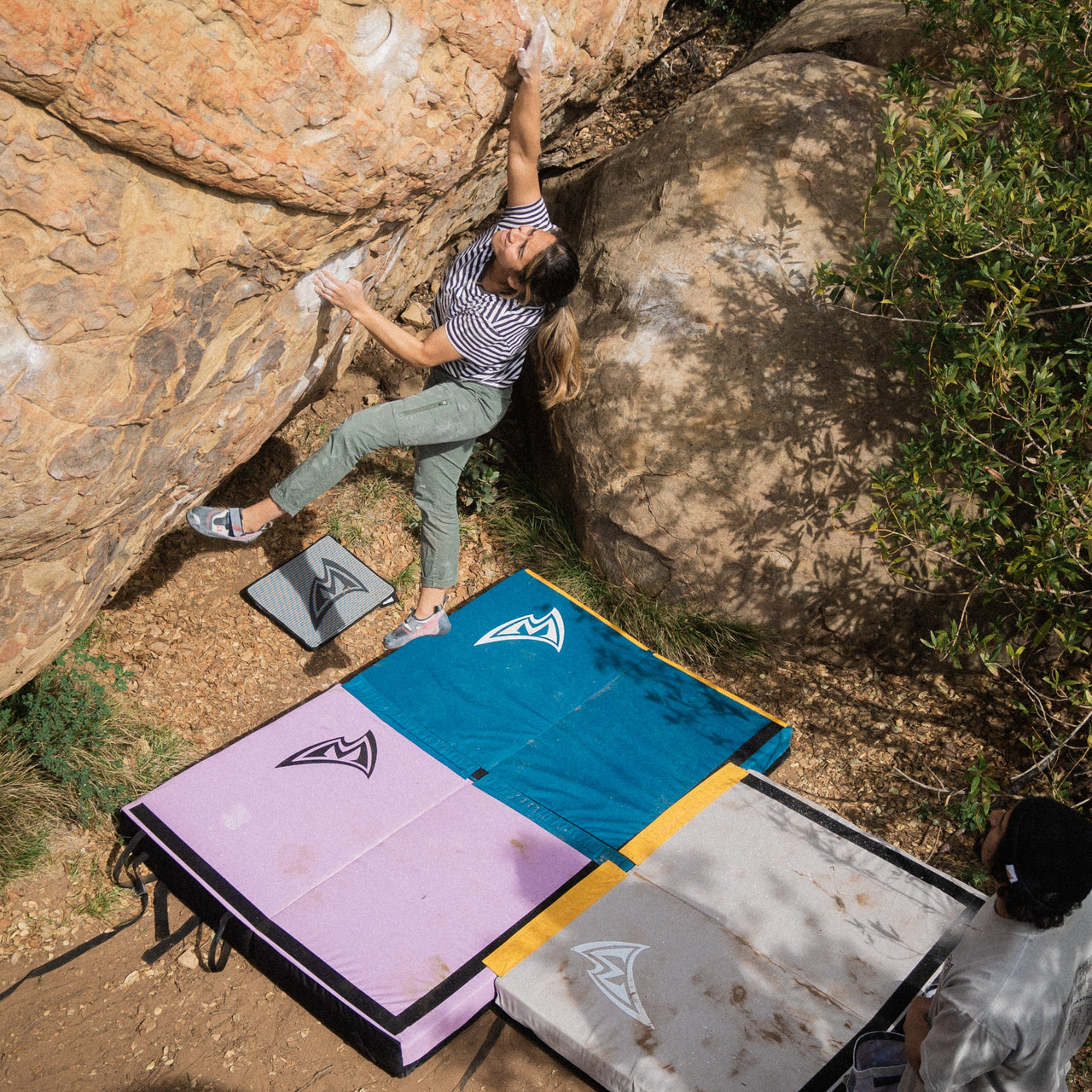 Person climbing a rock face with crash pads stitched together covering a wide landing zone below the climber